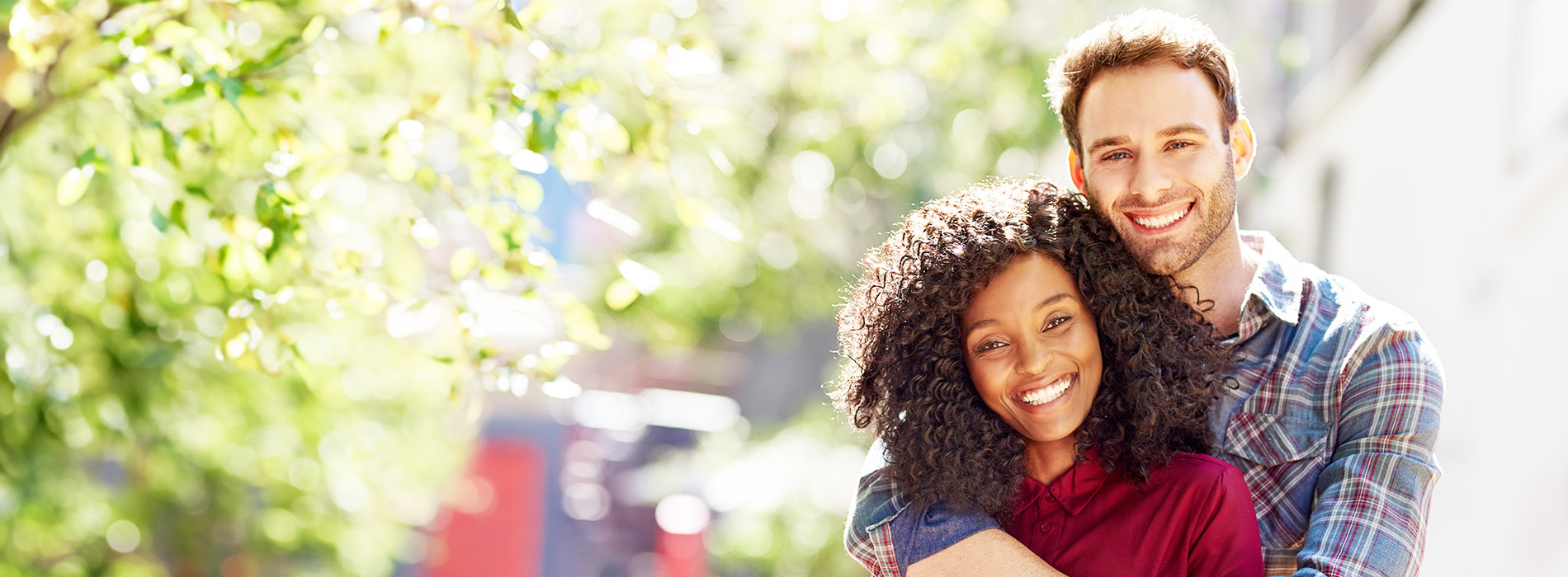 A man and a woman embrace in an outdoor setting, with the man wearing a plaid shirt and both smiling.