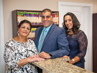 The image shows three individuals posing for a photo in an office setting. There is a man standing behind a counter, flanked by two women all are smiling and appear to be engaged in a professional environment.