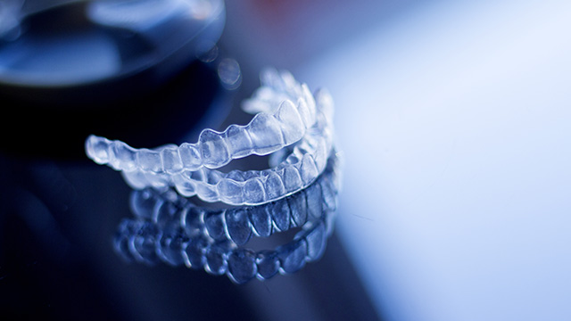 The image features a close-up of a transparent dental retainer with a watermark or logo visible, placed against a blurred background that suggests a domestic setting.