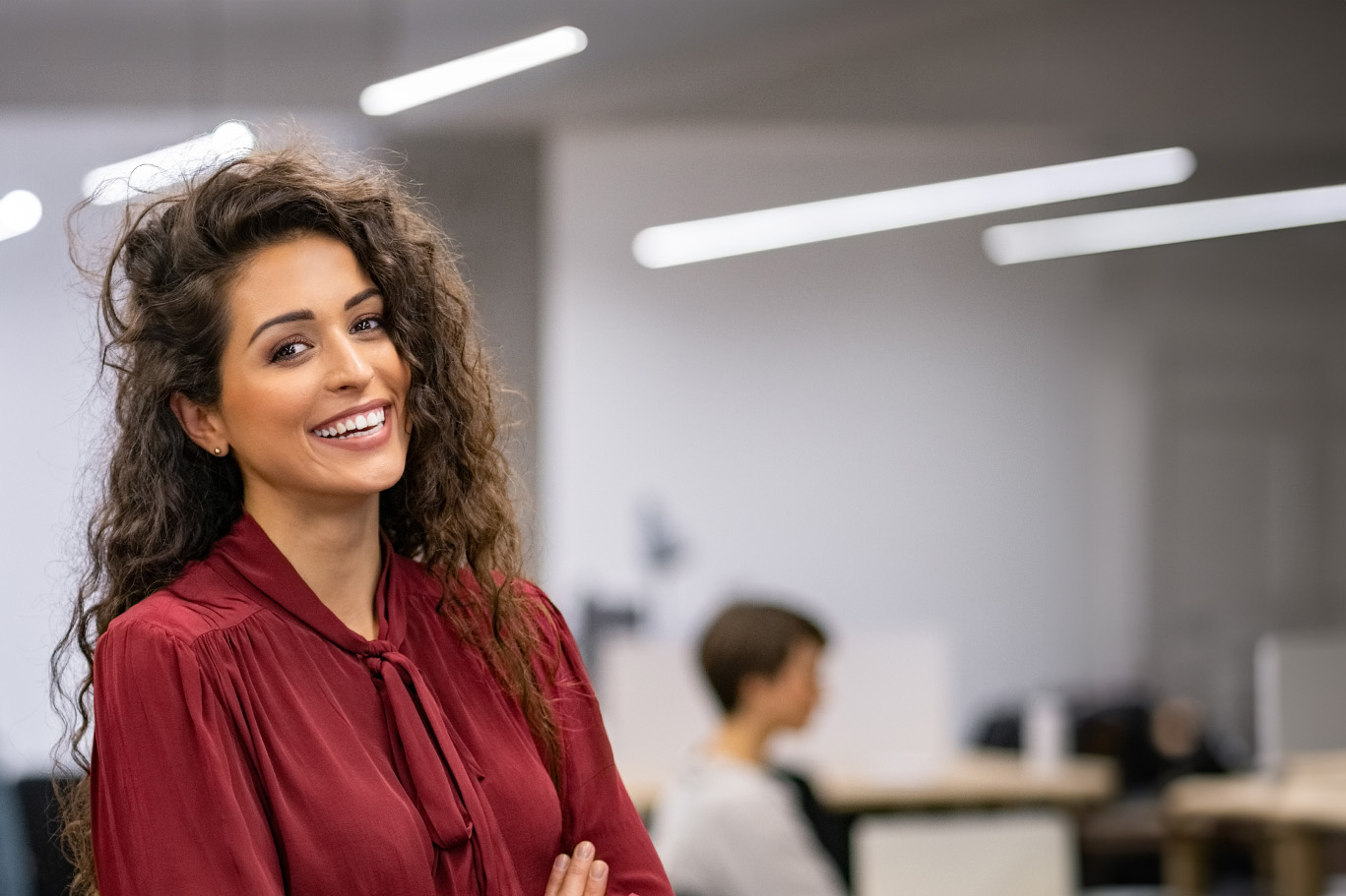 The image shows a smiling woman with curly hair, wearing a red top and standing in an office environment.